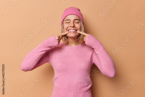 Cuadro en lienzo Happy millennial girl points at toothy smile keeps eyes closed shows perfect white teeth after whitening wears pink hat and jumper isolated over brown background feels positive and carefree