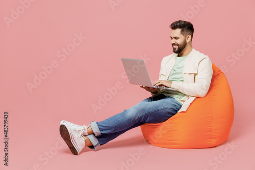 Full body young smiling happy man in trendy jacket shirt sit in bag chair hold use work on laptop pc computer isolated on plain pastel light pink background studio portrait. People lifestyle concept.