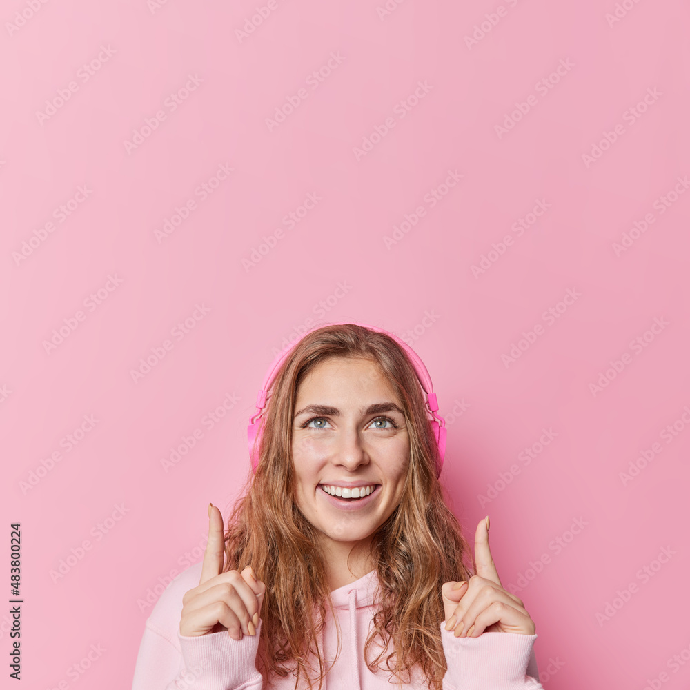 Pretty long haired woman smiles gladfully dressed in casual sweatshirt listens favorite melody points index fingers overhead isolated over pink background suggests to click on link. Look at this Pretty long haired woman smiles gladfully dressed in casual sweatshirt listens favorite melody points index fingers overhead isolated over pink background suggests to click on link. Look at this