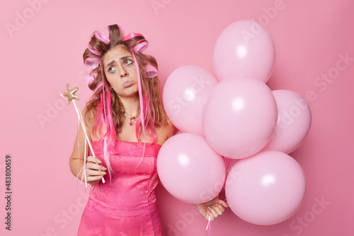 Frustrated unhappy European woman looks sadly away wears festive dress makesperfect hairstyle with rollers holds magic wand and inflated balloons has spoiled mood expresses negative emotions