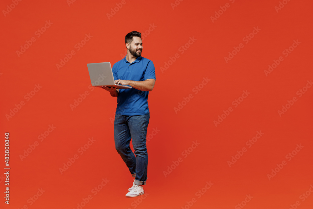 Full body young smiling happy caucasian man 20s in basic blue t-shirt looking aside on workspace area hold use work on laptop pc computer isolated on plain orange background People lifestyle concept Full body young smiling happy caucasian man 20s in basic blue t-shirt looking aside on workspace area hold use work on laptop pc computer isolated on plain orange background People lifestyle concept