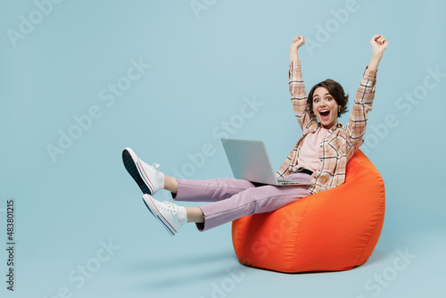 Full body young smiling happy woman 20s in brown shirt sit in bag chair with outstretched hands finish job hold use work on laptop pc computer isolated on pastel plain light blue background studio photo