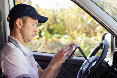 Focused deliveryman checking mobile phone in car. Side view of mid adult man in white T-shirt and cap looking at screen of phone. Work, delivery service, shipment concept