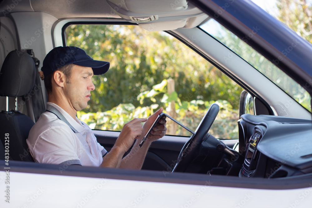 Side view of deliveryman checking mobile phone in car. Mid adult man in white T-shirt and cap looking at screen of phone. Using delivery app. Work, delivery service, shipment concept
