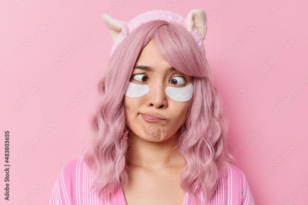 Horizontal shot of rosy haired Asian woman crosses eyes applies beauty patches for skin treatment has dyed pink hair wears soft headband poses against rosy background takes care of her complexion Horizontal shot of rosy haired Asian woman crosses eyes applies beauty patches for skin treatment has dyed pink hair wears soft headband poses against rosy background takes care of her complexion