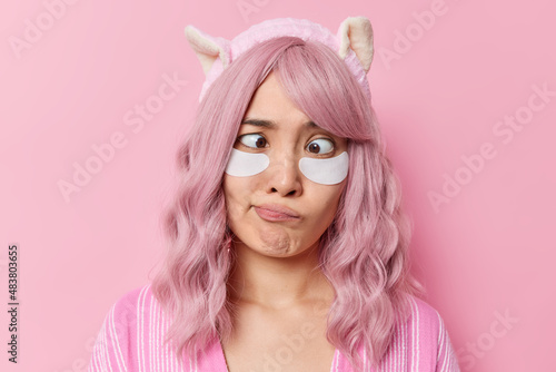 Horizontal shot of rosy haired Asian woman crosses eyes applies beauty patches for skin treatment has dyed pink hair wears soft headband poses against rosy background takes care of her complexion