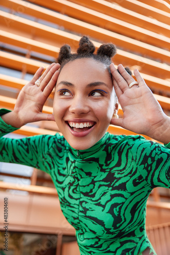 Vertical shot of happy young woman with bun hairstyle keeps pams near face smiles toothily applies blue eyeliner wears green jumper poses outdoors against blurred background has playful mood