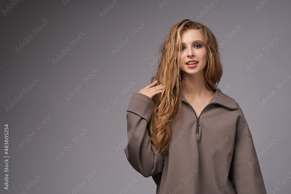 Happy blonde long haired woman model with makeup in grey oversize sweatshirt and yoga pants lifts up knee smiling in light studio closeup Happy blonde long haired woman model with makeup in grey oversize sweatshirt and yoga pants lifts up knee smiling in light studio closeup
