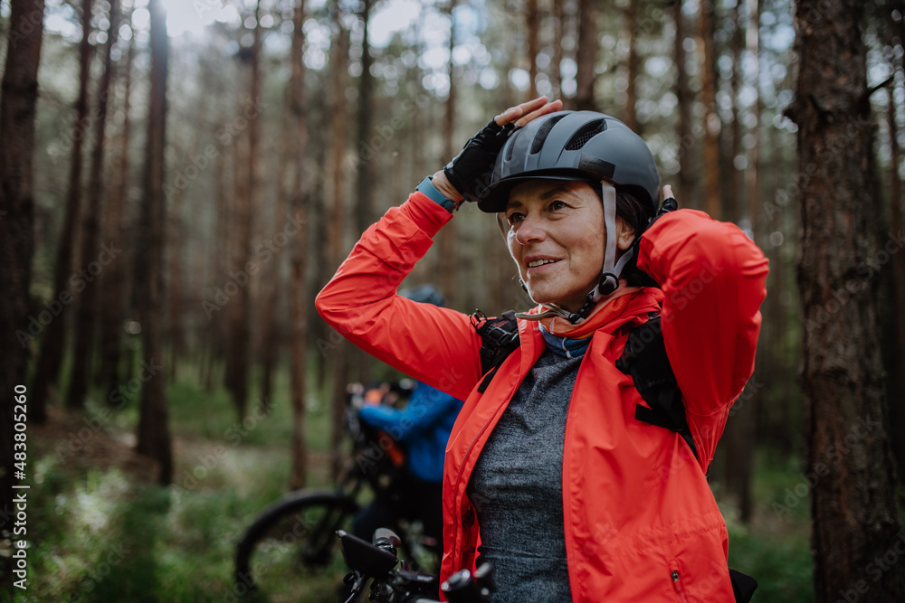 Senior woman biker putting on cycling helmet outdoors in forest in autumn day.