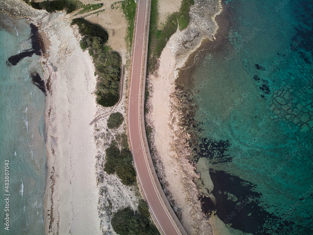 Aerial drone view of road passing trough a idyllic isthmus neck with sandy beach and vibrant blue sea, (Rena di ponente e levante) Sardinia.