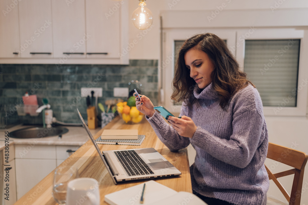 Hispanic woman with laptop using credit card and smartphone in kitchen at home Hispanic woman with laptop using credit card and smartphone in kitchen at home