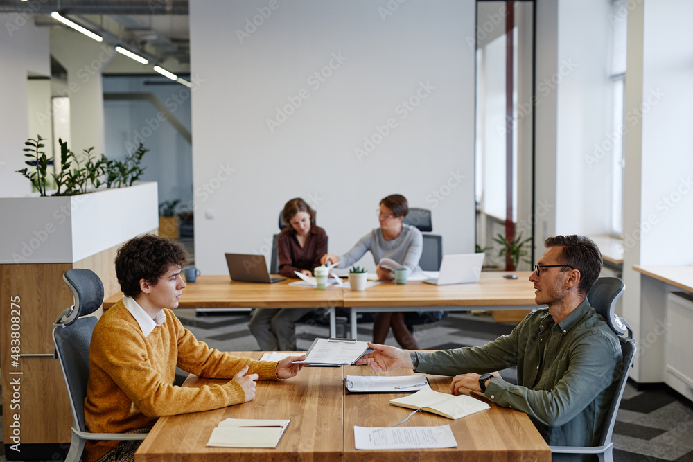 Side view portrait of HR manager handing contract to young man at job interview in office, copy space