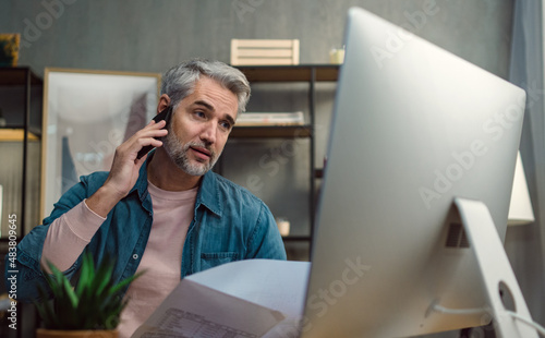 Mature man architect working on computer and making phone call at desk indoors in office.