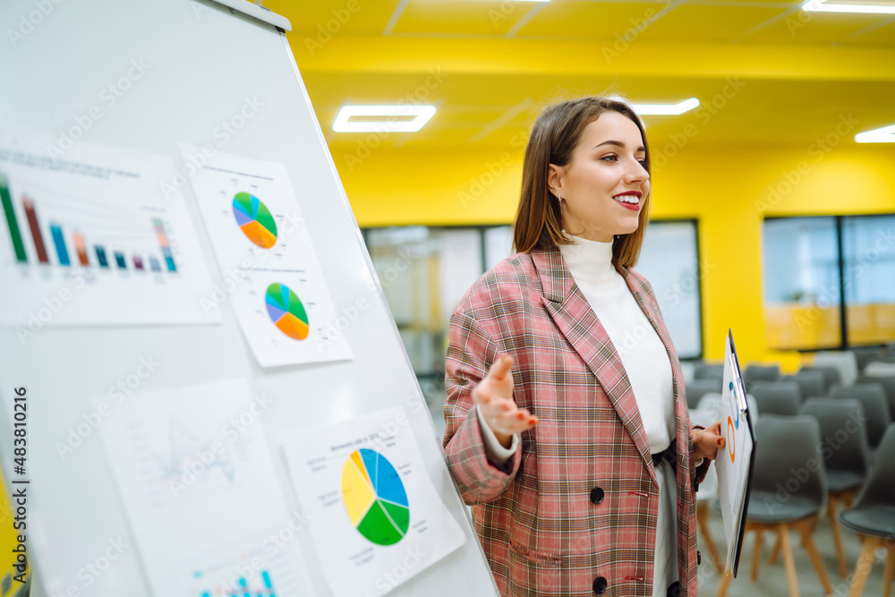 © maxbelchenko - Young woman leader, presenter, make a business presentation at a conference in the office, business woman mentor explaining a graphic diagram on a flipchart, corporate group seminar.