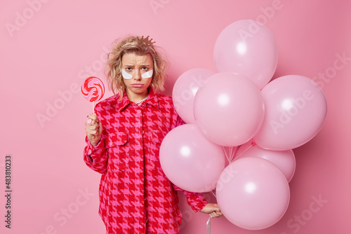 Frustrated upset woman has bad mood after party holds delcious round candy on stick and bunch of inflated airballoons wears beauty patches dressed in fashionable clothes isolated over pink wall