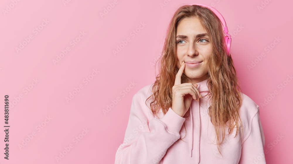 Smiling pretty young woman with long hair keeps finger near lips focused away thinks about something dressed in casual hoodie listens music via headphones isolated over pink backgroud copy space Smiling pretty young woman with long hair keeps finger near lips focused away thinks about something dressed in casual hoodie listens music via headphones isolated over pink backgroud copy space