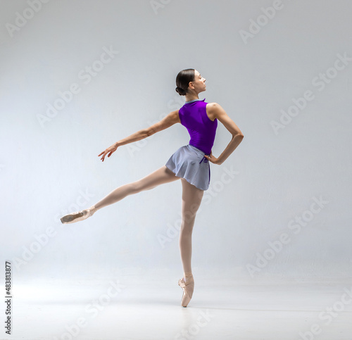 Fototapeta Naklejka Na Ścianę i Meble -  Young and beautiful ballerina in purple ballet leotard and grey skirt. She wears ballet pointe shoes and dancing in white studio.