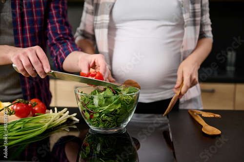 Focus on male hands holding a kitchen knife and puting sliced tomatoes into a bowl with lettuce leaves and greens on the background of his pregnant wife standing by a kitchen island.