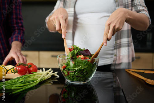 Focus on pregnant woman's hands holding wooden spoons and mixing ingredients in a glass bowl, preparing delicious healthy salad for dinner in the kitchen island next to her husband chopping vegetables
