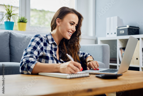 Female student writing notes using laptop while studying at home