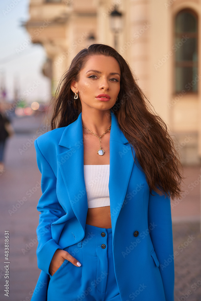 Confident young businesswoman with loose brown hair in blue pantsuit with handbag stands on city historical street at sunset in summer Confident young businesswoman with loose brown hair in blue pantsuit with handbag stands on city historical street at sunset in summer