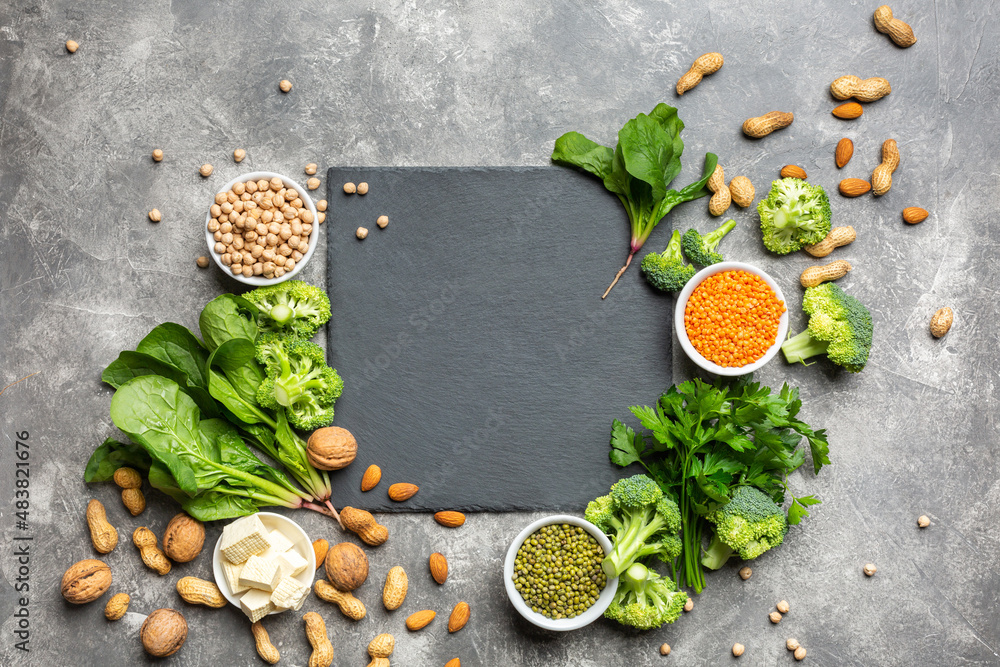 A source of protein for vegetarians. Healthy clean food: greens, vegetables, nuts and legumes top view on a concrete background with a black cutting stone in the center. A source of protein for vegetarians. Healthy clean food: greens, vegetables, nuts and legumes top view on a concrete background with a black cutting stone in the center.