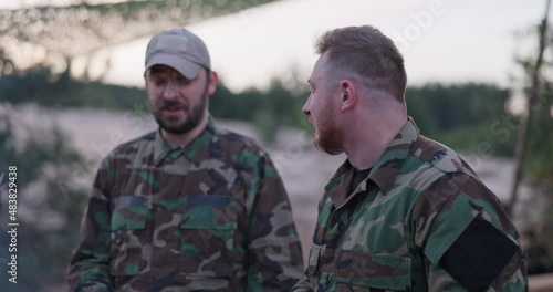 Soldiers in camouflage clothing, discussing in the evening at a common bonfire frying sausages for supper, men joking, laughing, resting after work, service, field exercises photo