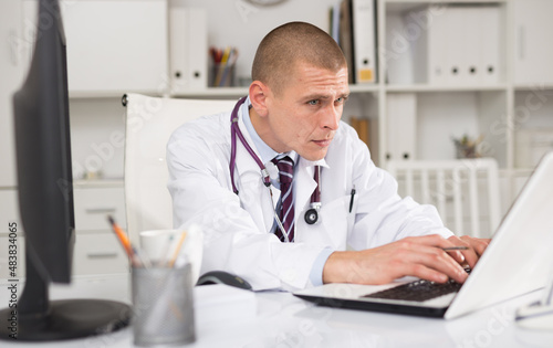 Portrait of european doctor sitting in white medical office and working on laptop computer