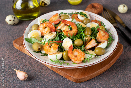 Salad with shrimps, quail eggs, olives and arugula on brown background