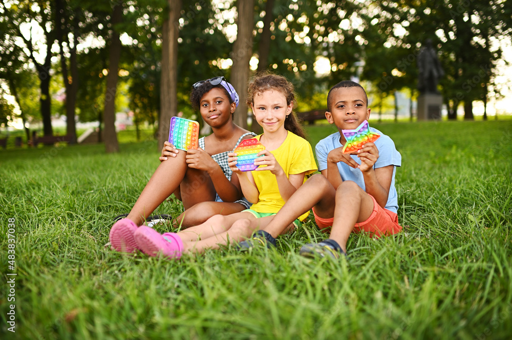 children of different races sit on the grass and play with an anti-stress toy pop-it children of different races sit on the grass and play with an anti-stress toy pop-it