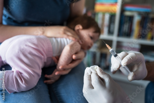 Fototapeta Naklejka Na Ścianę i Meble -  Close up on hand of unknown man doctor wearing protective medical gloves and holding a syringe preparing injection shot with vaccine for small caucasian baby mother hold her child for vaccination