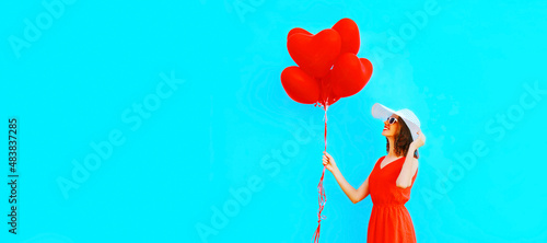 Beautiful happy smiling young woman with bunch of red heart shaped balloons wearing sunglasses on blue background