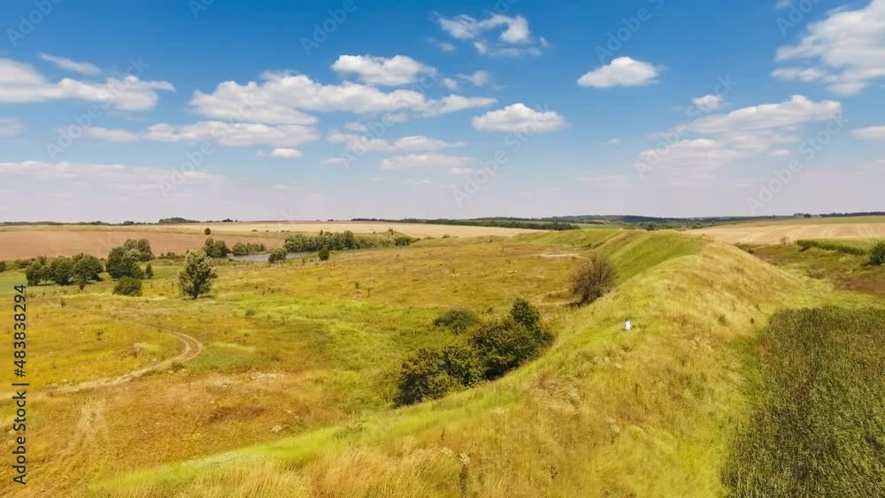 Woman in white dress walks along the hill. Sunny meadow bordering with farmlands. Bright blue sky with white clouds at backdrop. Woman in white dress walks along the hill. Sunny meadow bordering with farmlands. Bright blue sky with white clouds at backdrop.