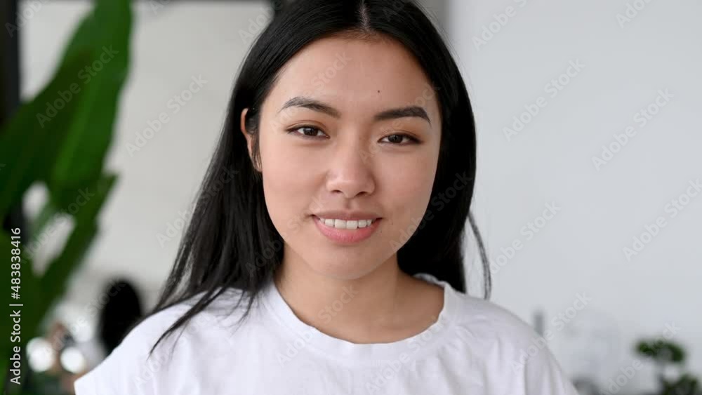 Close-up video portrait of a pretty young woman. Lovely happy girl of Asian ethnicity with long brunette hair, in a white t-shirt, stands at home, looking directly at the camera, smiling friendly Close-up video portrait of a pretty young woman. Lovely happy girl of Asian ethnicity with long brunette hair, in a white t-shirt, stands at home, looking directly at the camera, smiling friendly