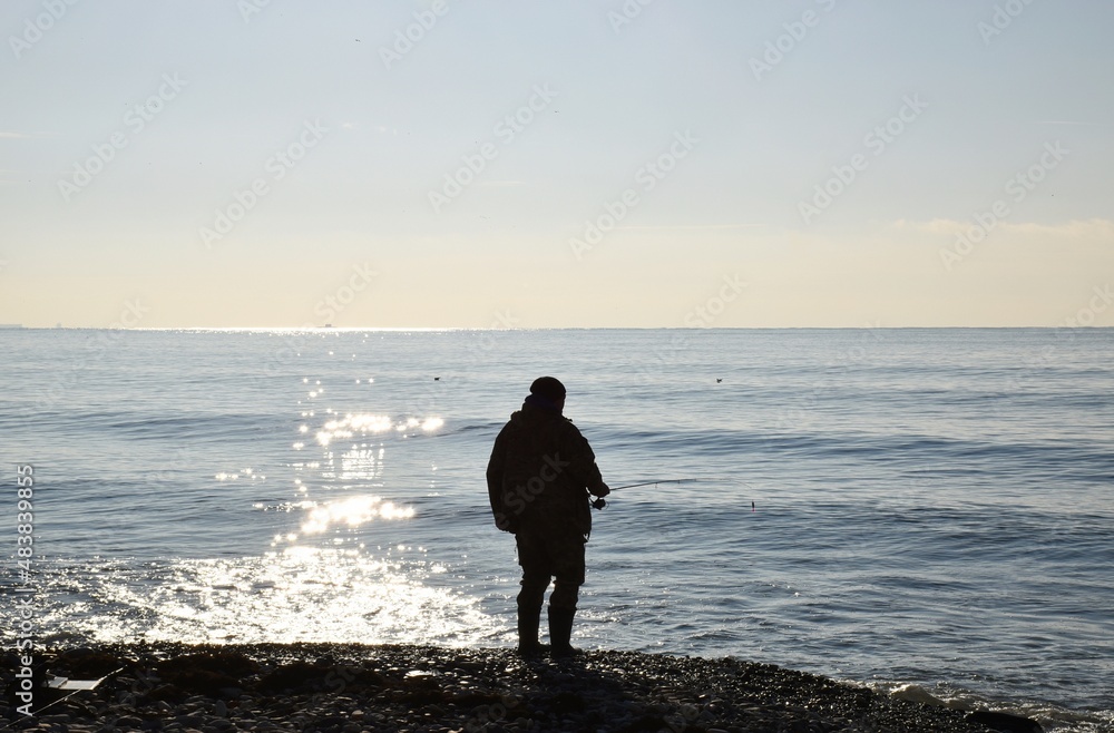 Calm morning sea. A fisherman on the shore catches fish.The sun's rays sparkle and run through the sea water. Calm morning sea. A fisherman on the shore catches fish.The sun's rays sparkle and run through the sea water.