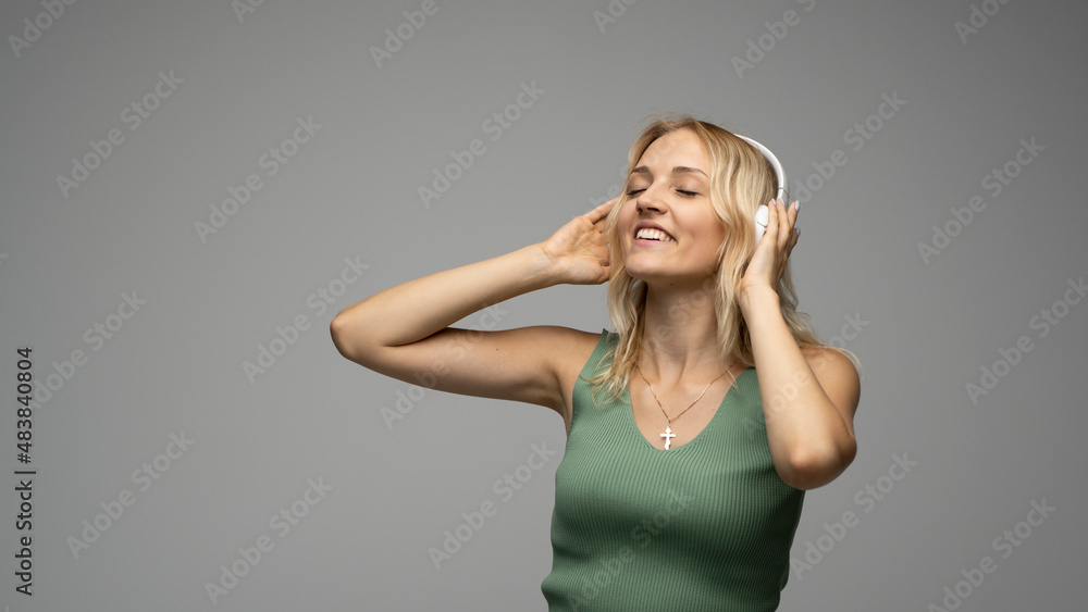 Beautiful attractive young blond woman wearing green t-shirt in white headphones listening music on grey background in studio. Lifestyle. Beautiful attractive young blond woman wearing green t-shirt in white headphones listening music on grey background in studio. Lifestyle.