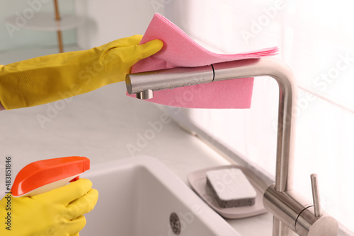 Woman cleaning faucet with rag and detergent in kitchen, closeup