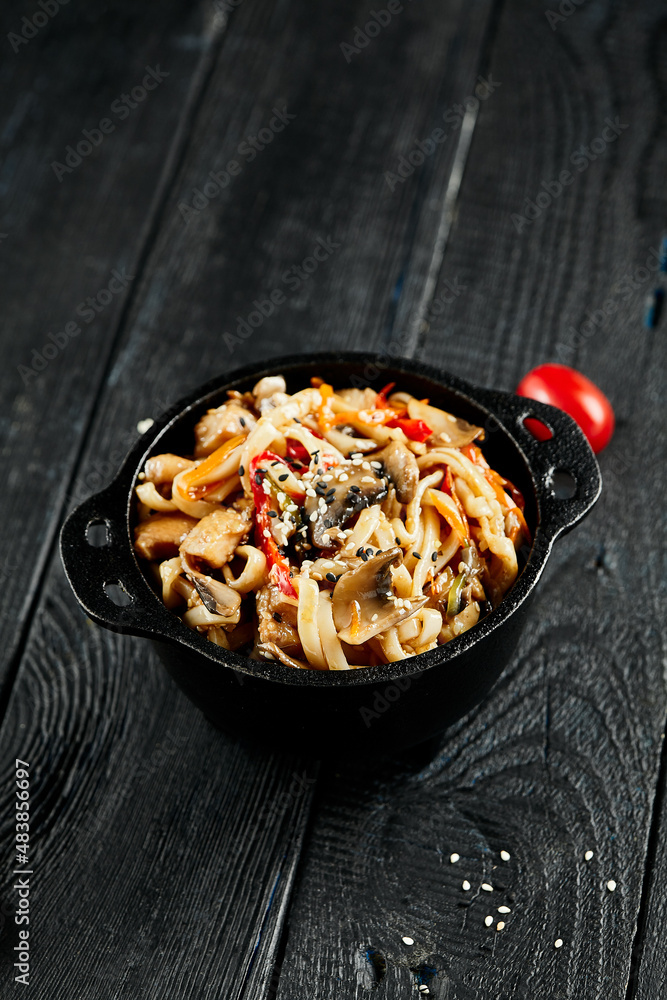 Stir fried udon noodles in wok. Udon noodle with chicken and mushrooms on wooden background. Asian noodles on black table with ingredients. Wok menu for japanese restaurant. Stir fried udon noodles in wok. Udon noodle with chicken and mushrooms on wooden background. Asian noodles on black table with ingredients. Wok menu for japanese restaurant.
