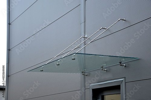 Glass roof above the entrance to the industrial building with metal cladding of the facade. thick-walled glass panel hangs above the door on metal rods with round targets holders