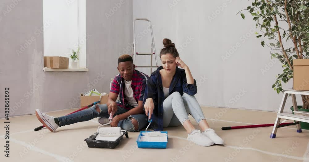 A couple of lovers are sitting on the floor in a renovated apartment, they are applying paint poured in paints trays on rollers, talking about decorations, finishing the house A couple of lovers are sitting on the floor in a renovated apartment, they are applying paint poured in paints trays on rollers, talking about decorations, finishing the house
