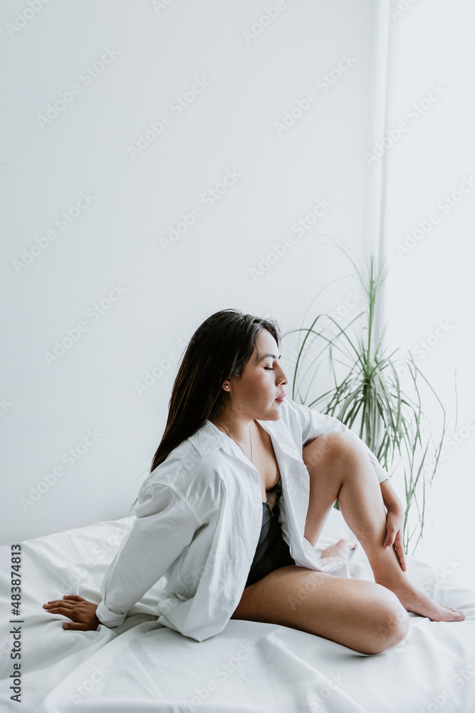 Young Latin woman sitting on bed, wearing white robe, showing legs at home in Mexico city Young Latin woman sitting on bed, wearing white robe, showing legs at home in Mexico city