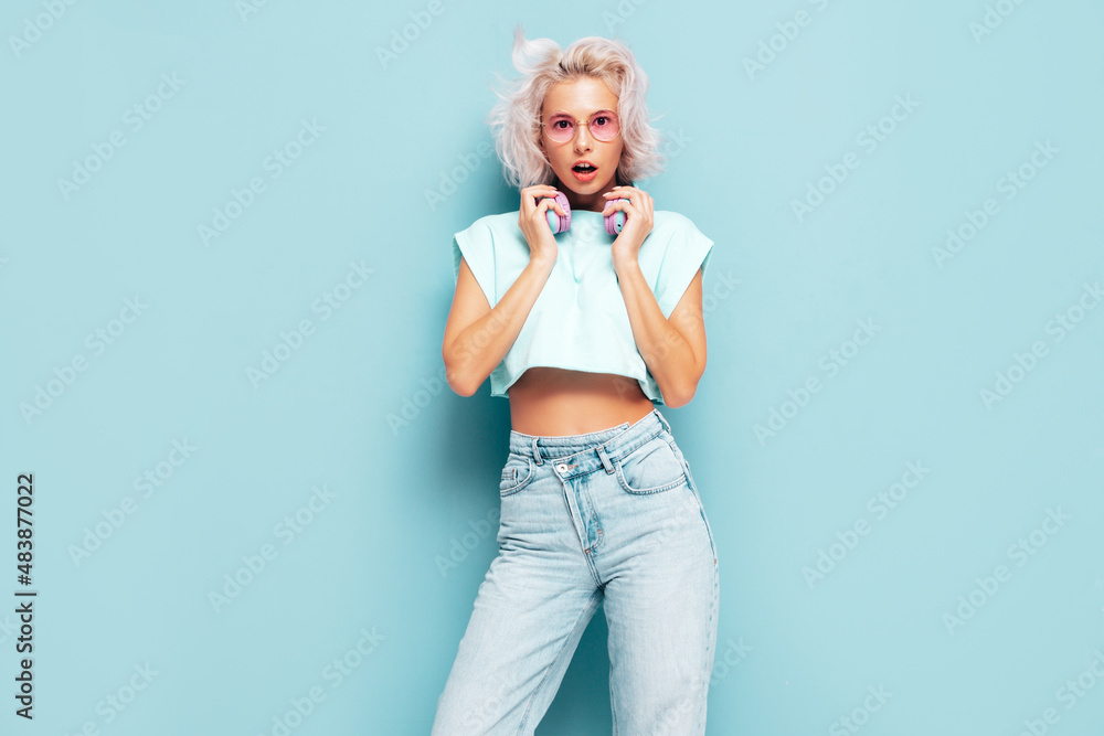 Young beautiful smiling female in trendy summer clothes. Sexy carefree woman posing near blue wall in studio. Positive blond model having fun. Listening music in wireless headphones Young beautiful smiling female in trendy summer clothes. Sexy carefree woman posing near blue wall in studio. Positive blond model having fun. Listening music in wireless headphones