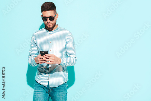 Handsome model.Sexy stylish man dressed in shirt and jeans. Fashion hipster male posing near blue wall in studio. Holding smartphone. Looking at cellphone screen. Using apps. In sunglasses