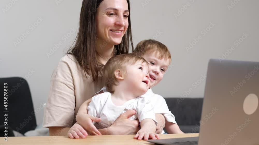 Happy and tired mother working from home, working at the table with a laptop, two sons are sitting in her arms. The concept of motherhood and parenting. Happy and tired mother working from home, working at the table with a laptop, two sons are sitting in her arms. The concept of motherhood and parenting.