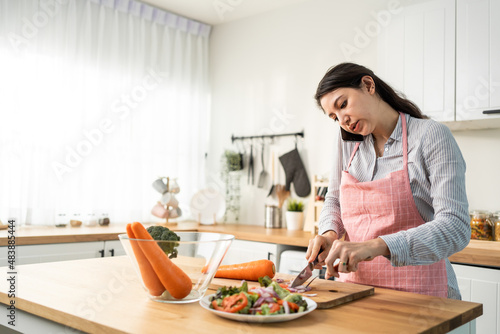 Caucasian woman talk on phone while cooking salad in kitchen at home. 