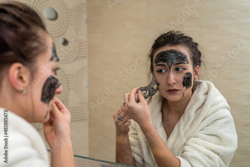 Fototapeta Naklejka Na Ścianę i Meble -  young cute brunette makes a gorgeous clay black mask from pimples and blackheads.