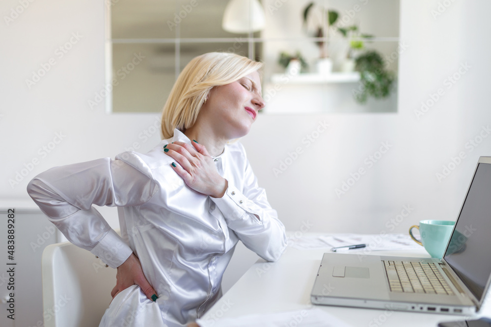 Portrait of young stressed woman sitting at home office desk in front of laptop, touching aching neck with pained expression, suffering from neck pain after working on laptop Portrait of young stressed woman sitting at home office desk in front of laptop, touching aching neck with pained expression, suffering from neck pain after working on laptop