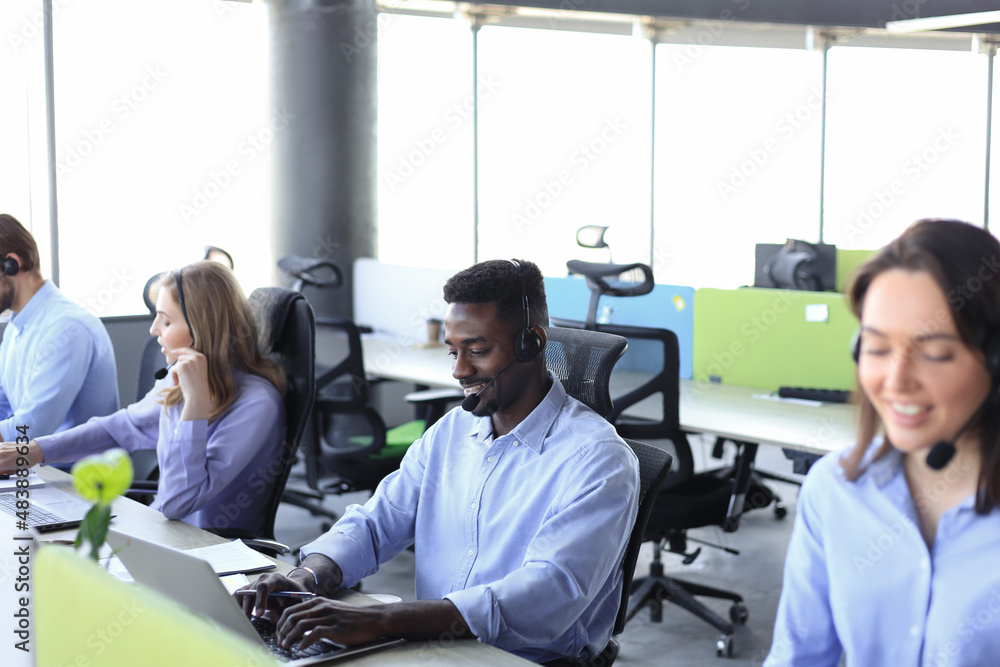 Obraz premium Smiling african male call-center operator with headphones sitting at modern office with collegues on the backgroung, consulting online.