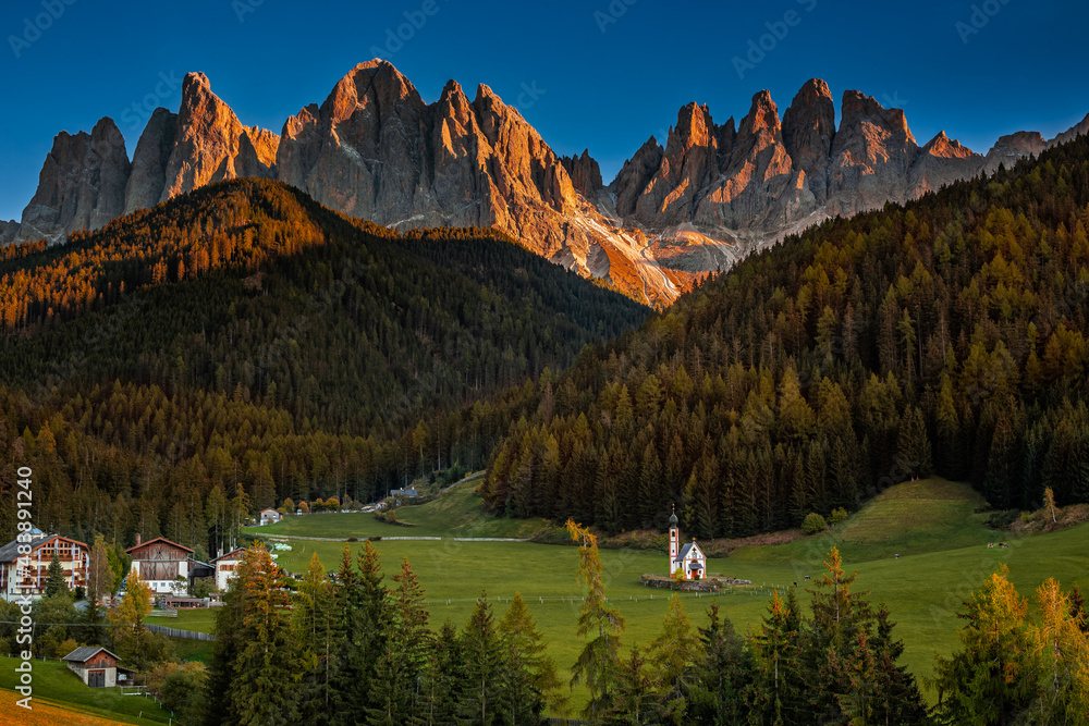 Val Di Funes, Dolomites, Italy - Aerial view of the beautiful St. Johann Church (Chiesetta di San Giovanni in Ranui) at South Tyrol with the Italian Dolomites in warm sunset at background and blue sky