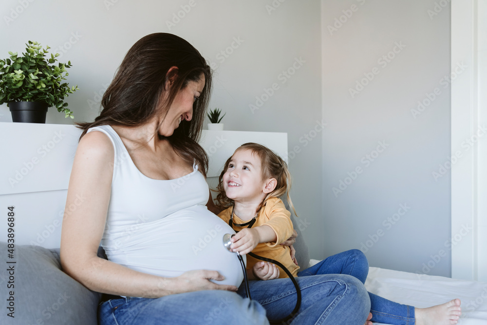 Smiling girl with stethoscope looking at pregnant mother sitting on bed Smiling girl with stethoscope looking at pregnant mother sitting on bed